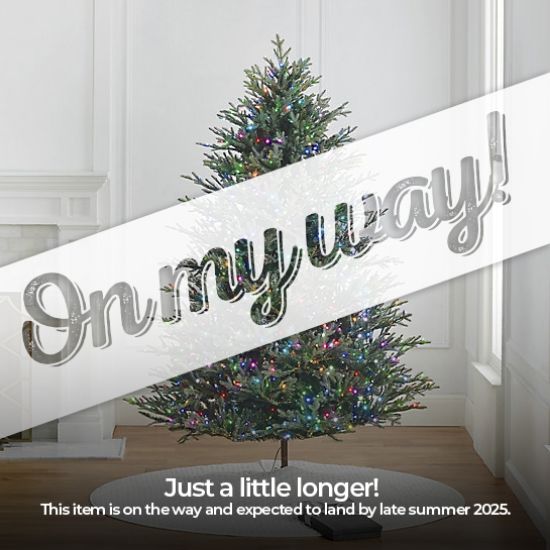 7.5' Full Fraser Pine displayed in lifestyle shot of living room, lit multi-color, graphic banner across the image with text saying "On my way!". Caption under banner says "Just a little longer! This item is on the way and expected to land by late summer 2025."